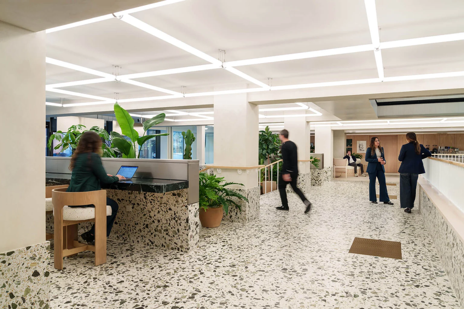 People working and walking through the bright ground-floor lounge, with terrazzo floors and planting